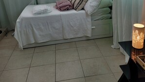 Un lit bien fait, avec du linge de lit blanc et une couverture rose, une table de chevet avec une lampe, et un mur vert.