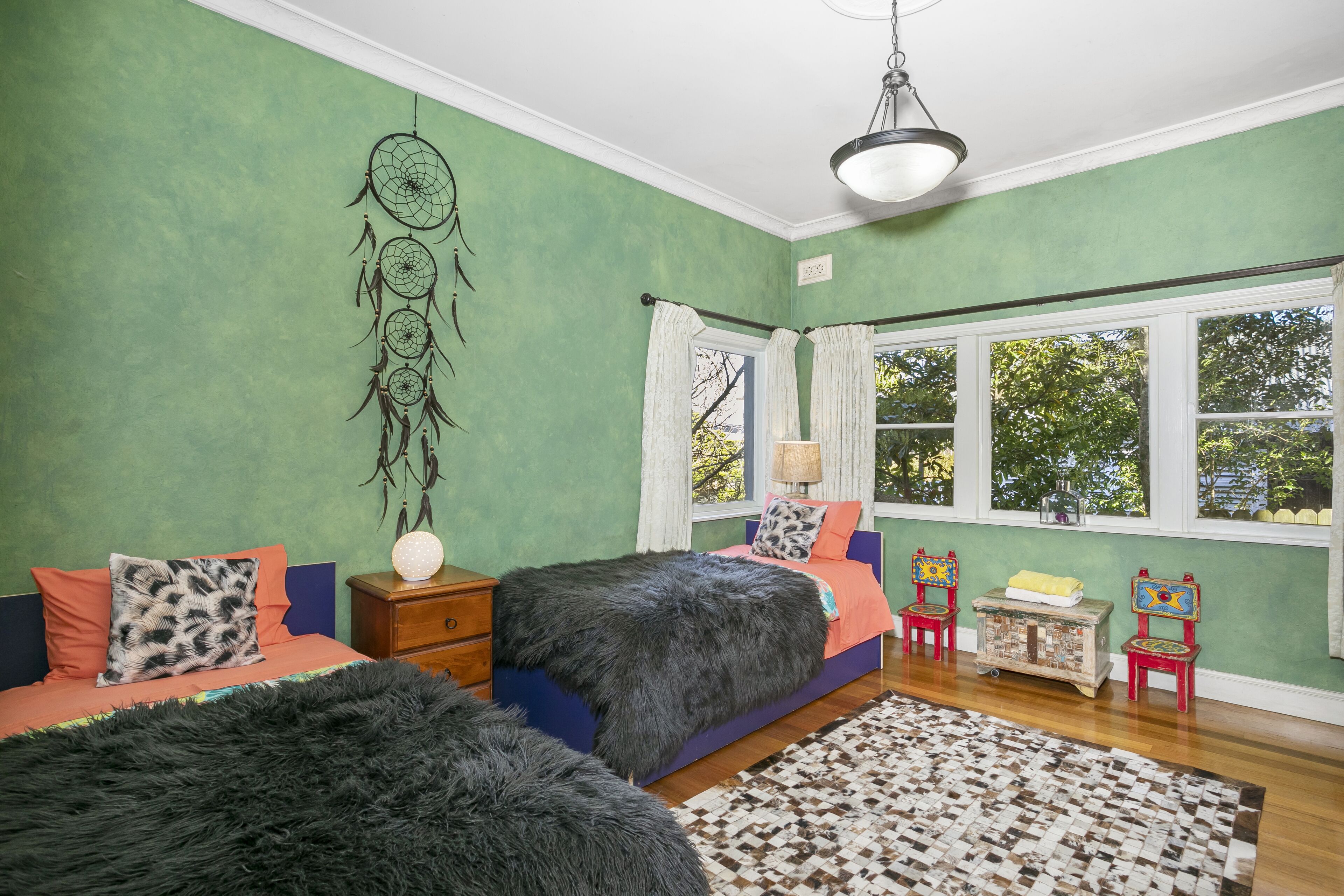 A bedroom with two beds, a dreamcatcher, a window with curtains, and a view of trees.