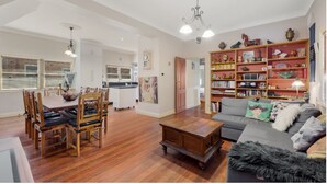 A spacious living room with a dining area, a kitchen in the background, and a large bookshelf filled with books and decorative items. - Welcome to our charming, 2-level house just 5 min walk to 3 Sisters, Echo Point! (Katoomba)