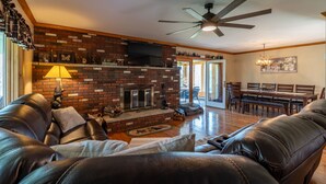 A living room with a brick fireplace, leather sofas, a dining area, and a ceiling fan. - Spruce Haven - 5 br home on 53 acres with pond (Meshoppen)