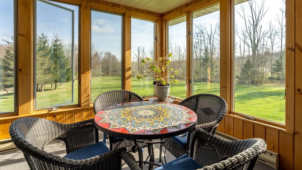 A covered patio with a round table and wicker chairs, large windows offering a view of a green landscape. - Spruce Haven - 5 br home on 53 acres with pond (Meshoppen)