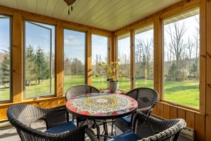 A covered patio with a round table and wicker chairs, large windows offering a view of a green landscape.