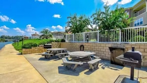 An outdoor patio with picnic tables and a grill, surrounded by a white fence and greenery. - Lovely Lake Condo with BOAT SLIP and Pool (Alexander City)