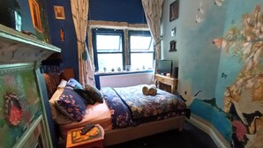 Une chambre à coucher comprenant un lit, une petite table, une commode, une fenêtre avec des rideaux et un mur recouvert d’un papier peint à motifs floraux.