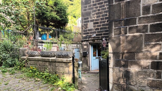 Un étroit sentier pavé de pierres menant à un bâtiment traditionnel en pierre, doté d’une porte bleue, et entouré de verdure et d’une clôture en métal.