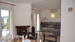 A dining area with a wooden table set for four, a sofa in the background, and a balcony with a view of trees. - Casa Gaudir - L'Estartit, Torre Vella (Estartit (L´))