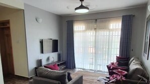 Una sala de estar con un sofá gris, una mesita pequeña y un televisor. Hay un ventilador de techo, una ventana con cortinas y una puerta que da a otra habitación.