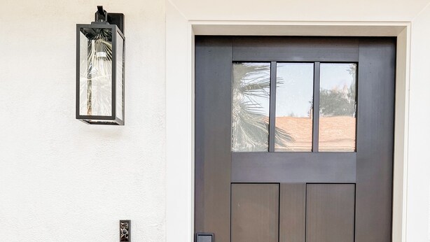A dark-colored front door with a glass panel, a wall-mounted light fixture, and a small electronic device on the wall. - Cozy Desert Remodeled 2-bedroom Home (Gilbert)