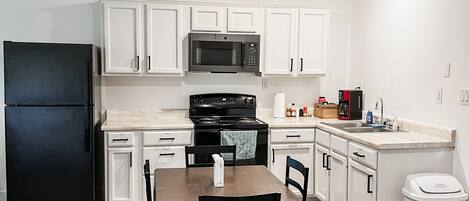 A compact kitchen with white cabinets, a black refrigerator, a microwave, a stove, and a dining table with chairs.