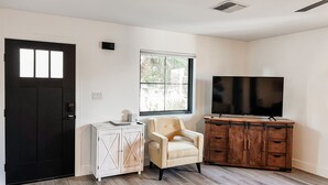 A room with a black door, a white cabinet with drawers, a beige armchair, a wooden cabinet with a TV, and a window with a view of greenery. - Cozy Desert Remodeled 2-bedroom Home (Gilbert)