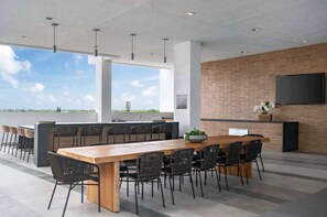 Un espace repas moderne avec une longue table en bois et des chaises noires, un mur de briques sur lequel est fixé un téléviseur, et de grandes fenêtres offrant une vue sur le ciel et le paysage.