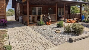 En rustik træhytte med en overdækket veranda, en grusvej og en lille have med planter og græskar.