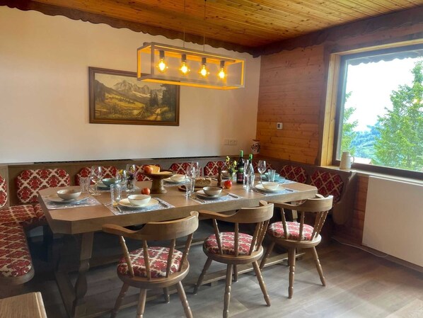 Un coin repas avec une table en bois dressée pour quatre personnes, des bancs dotés de coussins à motifs, un tableau accroché au mur et une fenêtre donnant sur la forêt.