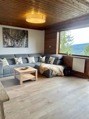 Un salon moderne avec un canapé d’angle gris, une table basse en bois et une grande fenêtre donnant sur l’extérieur.