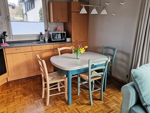 Une cuisine avec des meubles en bois, un four à micro-ondes, une table  à manger avec des chaises et une fenêtre avec des stores.