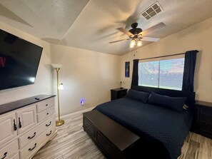 Slaapkamer met een bed, televisie, plafondventilator, vloerlamp, nachtkastje en een raam met jaloezieën.