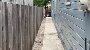 Un estrecho sendero de hormigón entre una valla de madera y una casa gris con antena parabólica y contadores.