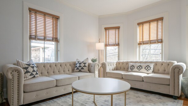 Una sala de estar con dos sofás color beige, una mesa de centro redonda blanca y persianas de madera en las ventanas.
