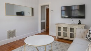 Una sala de estar con una mesa redonda blanca, un sofá blanco, un mueble de madera, un espejo grande y un televisor de pantalla plana montado en la pared.