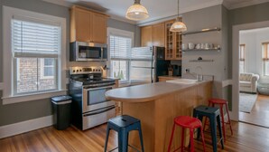 Una cocina con armarios de madera, electrodomésticos de acero inoxidable, una isla central con taburetes y vista al área de estar a través de una puerta abierta.