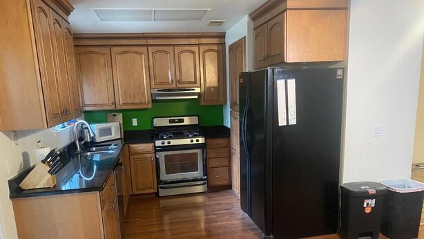 A kitchen with wooden cabinets, a black refrigerator, a microwave, a stove, and a trash bin.