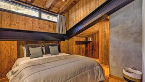 Ein gemütliches Schlafzimmer mit Holzbalkendecke, einem großen Bett mit grauer Bettdecke und einem Fenster mit Vorhängen.