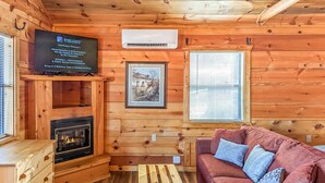 L’intérieur chaleureux d’un chalet, avec un décor en bois, une cheminée, un canapé rouge agrémenté de coussins, un téléviseur fixé au mur et un tableau encadré accroché au mur.
