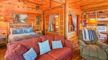 A cozy log cabin interior with a bed, red sofa, and armchair, wooden walls, and a kitchen area.