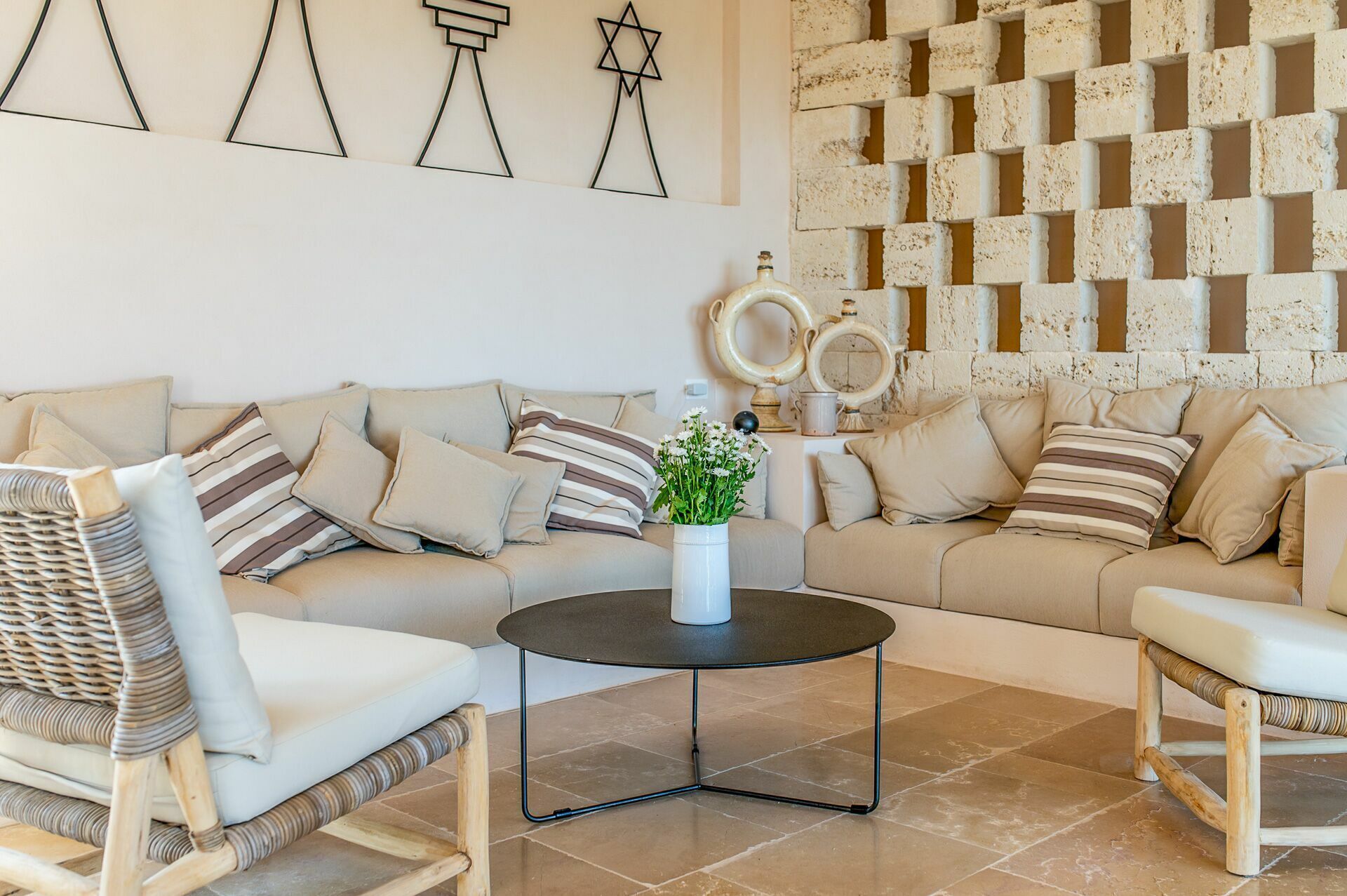 Un salon avec un canapé beige, des coussins à rayures, un fauteuil en osier, une table basse ronde et un mur orné d’un motif en briques et de divers objets décoratifs.