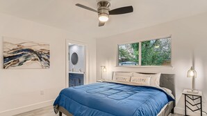 A bedroom with a blue bedspread, a ceiling fan, a window with a view of greenery, and a painting on the wall. - Beautiful Fully Remodeled Home (Austin)