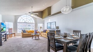 A spacious living area with a dining table set for six, a sofa, and a large window with blinds.