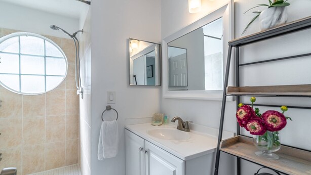 A bathroom with a shower, a sink, a mirror, a towel rack, and a shelf with flowers.