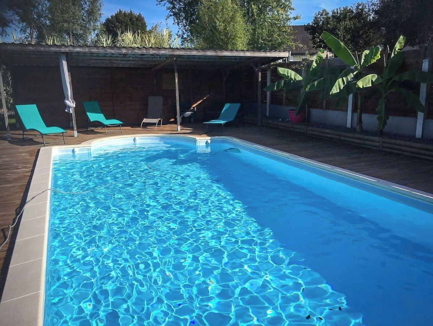 Une piscine rectangulaire à l’eau d’un bleu cristallin, entourée d’une terrasse en bois et de chaises longues vertes.