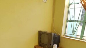 A small room with a television on a wooden shelf, a blue plastic chair, and a window with a metal grid. - Jaybee's Resort (Eldoret)