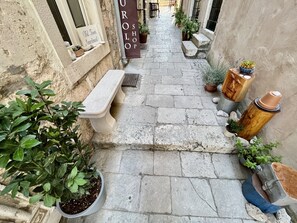 Une ruelle étroite, pavée de pierres, avec un banc, des plantes en pot et une enseigne indiquant « Appartement dans le vieux quartier ».