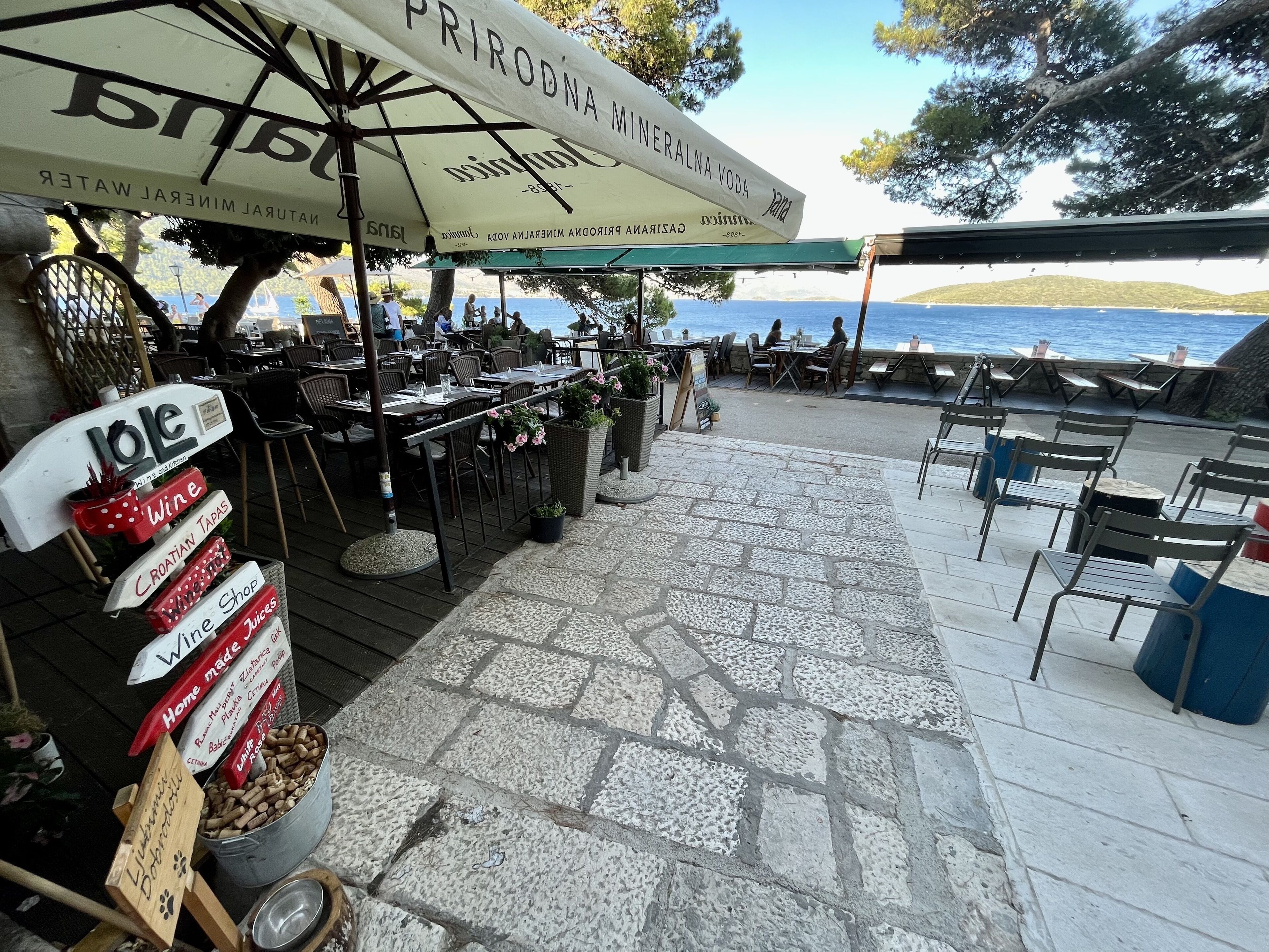 An outdoor dining area with tables and chairs, a sign for a mineral water brand, and a sign for a Croatian wine shop.