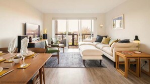 A modern living room with a sofa, dining table, and chairs. There is a TV mounted on the wall and a window providing natural light. - Rampart View -  a house that sleeps 6 guests  in 3 bedrooms (Gosport)