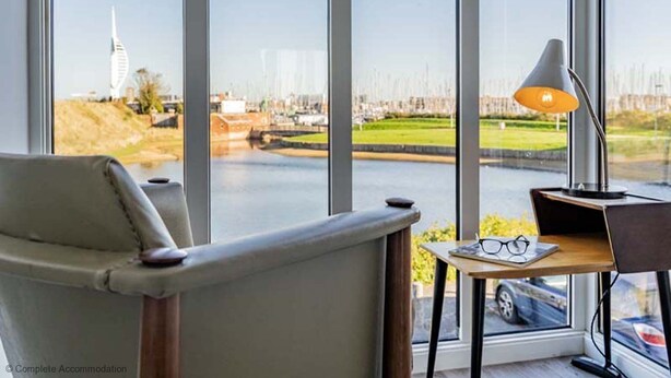 A modern room with a large window, a chair, a desk with a lamp, and a pair of glasses on the desk. - Rampart View -  a house that sleeps 6 guests  in 3 bedrooms (Gosport)