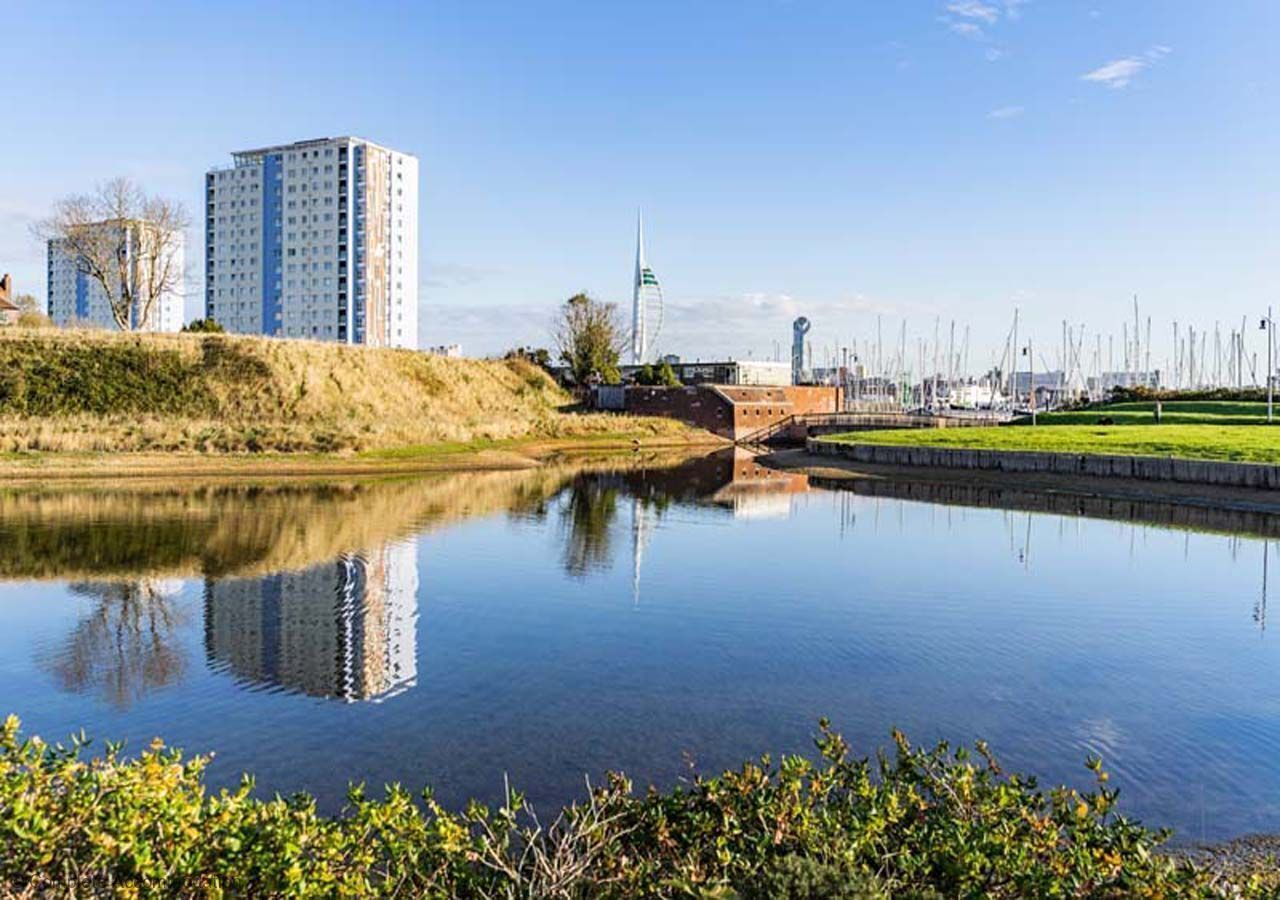 A calm river reflecting buildings, including a tall residential tower and a wind turbine.