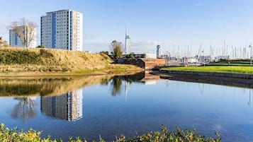 A calm river reflecting buildings, including a tall residential tower and a wind turbine.