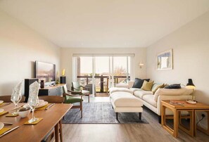 A modern living room with a sofa, dining table, and chairs. There is a TV mounted on the wall and a window providing natural light. - Rampart View -  a house that sleeps 6 guests  in 3 bedrooms (Gosport)