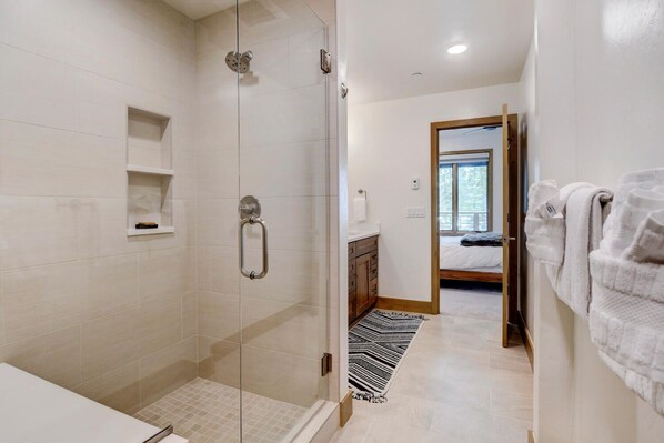 A modern bathroom with a glass shower enclosure, a wooden vanity, and a black and white striped rug leading to a bedroom with a bed and a window.