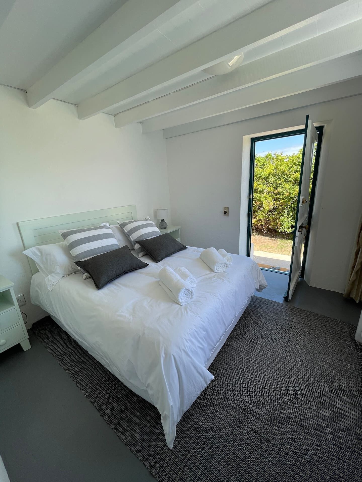 Un dormitorio con cama, almohadas, una mesita de noche y vistas a la vegetación a través de una puerta abierta.