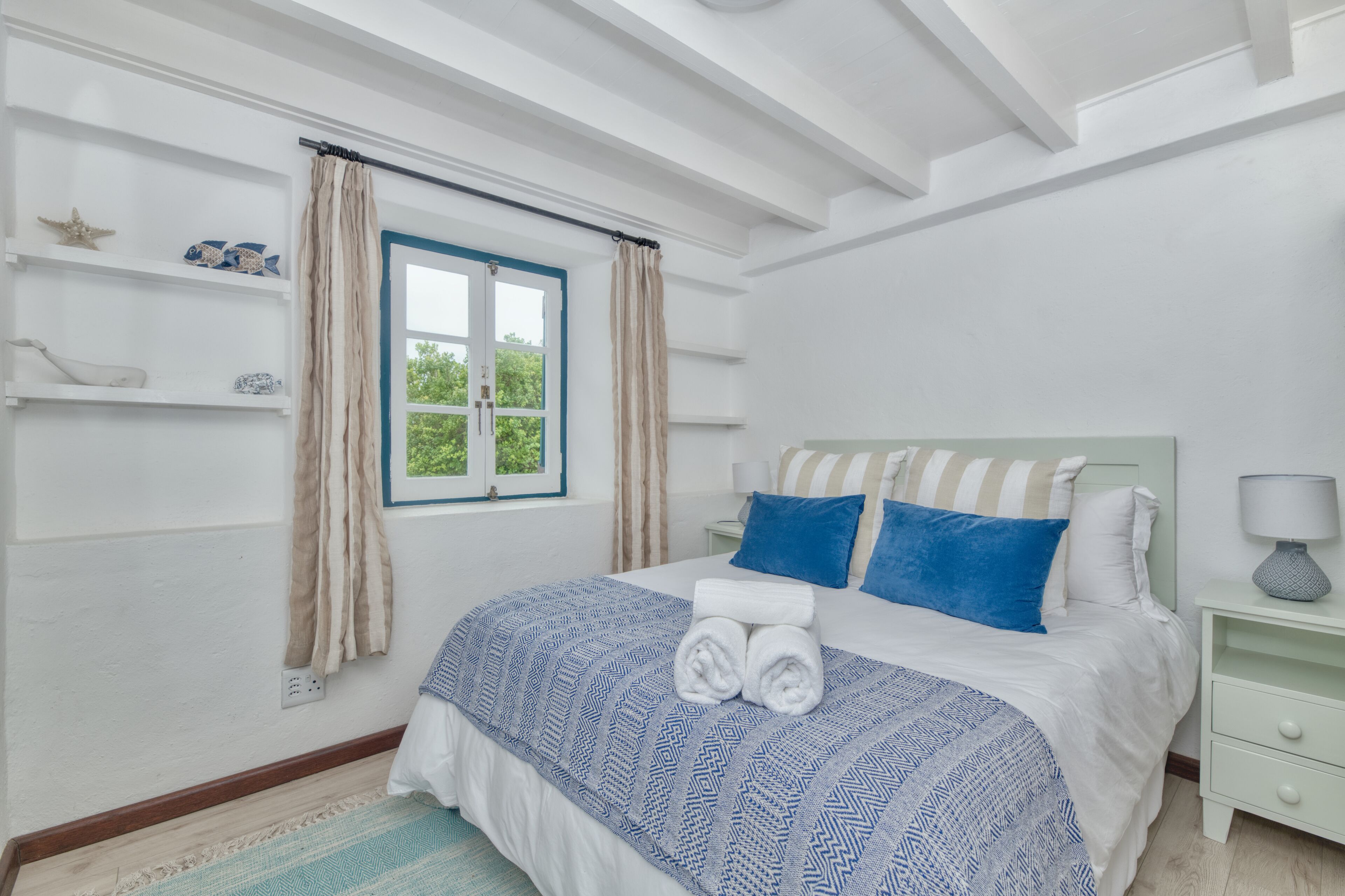 Un dormitorio con cama, mesita de noche, ventana con cortinas y estante con objetos decorativos.
