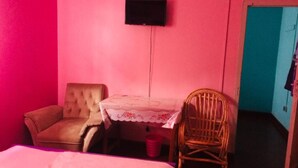 Ein Zimmer mit rosa Wänden, einem Einzelbett, einem hölzernen Stuhl, einem kleinen Tisch mit Spitze und einem an der Wand befestigten Fernseher.