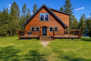 Une cabane en bois à deux étages, dotée d’une terrasse à l’avant, d’une entrée centrale et d’une aire de jeux à l’arrière-plan.