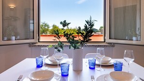 Ein Esstisch ist gedeckt mit blauem Glas, weißen Tellern und einer weißen Vase mit Grünpflanzen. Im Hintergrund sind Bäume und ein Gebäude zu sehen.