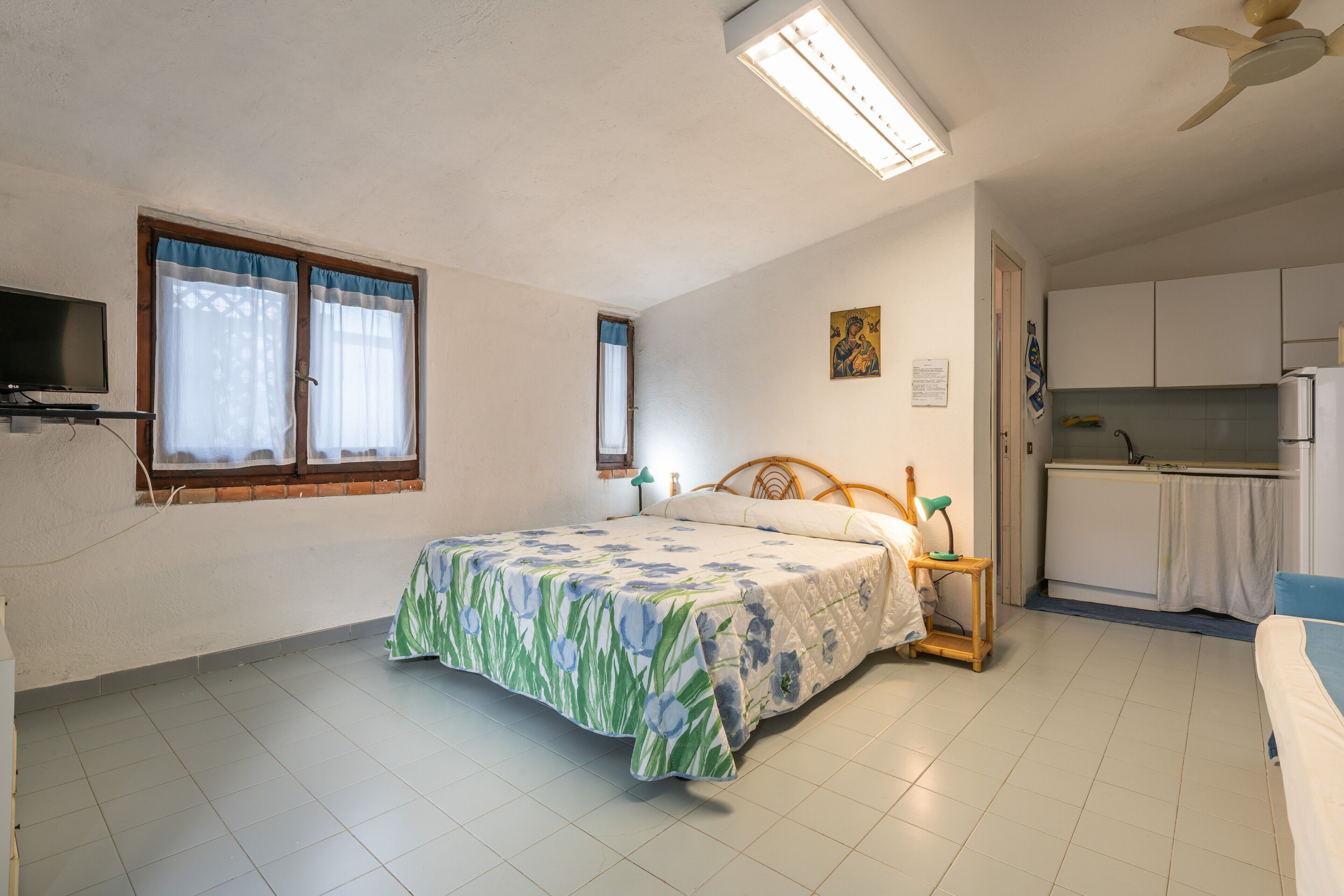 A bedroom with a bed, a desk, a chair, a window, and a kitchenette.