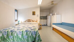 Une chambre à coucher avec un lit, une table de chevet, une lampe, une fenêtre, un ventilateur de plafond et, au fond, une petite cuisine.