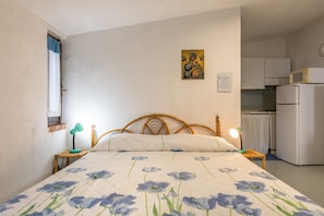 A bedroom with a bed, a nightstand, a lamp, a wall with a religious icon, and a kitchen area.